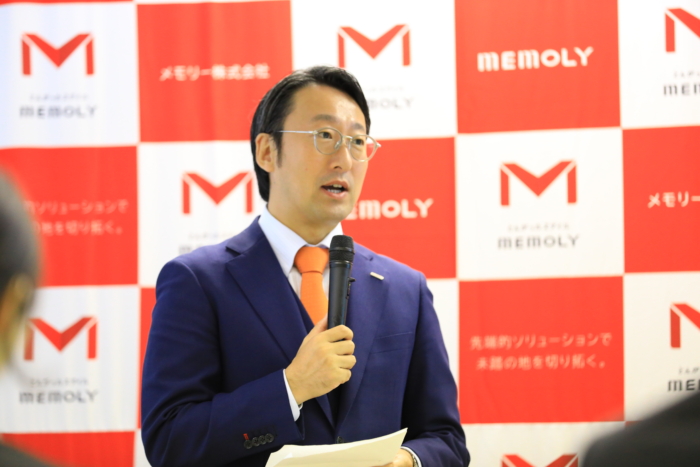 赤い弾幕の前で訓辞を話すスーツの男性