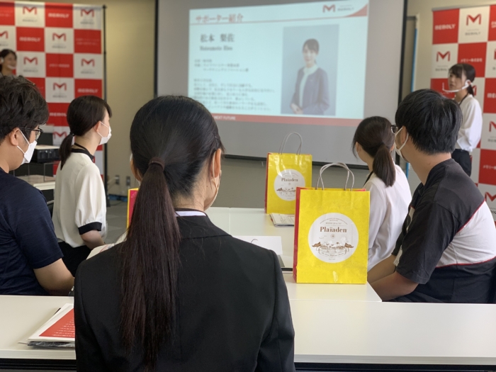 スクリーンの前で自己紹介をする社員の話を聞く学生