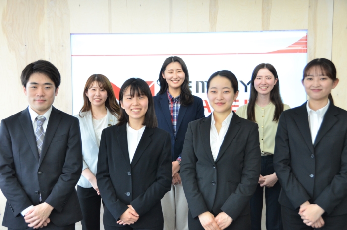 屋内スライドの前でスーツを着た7人が集合写真を撮っている