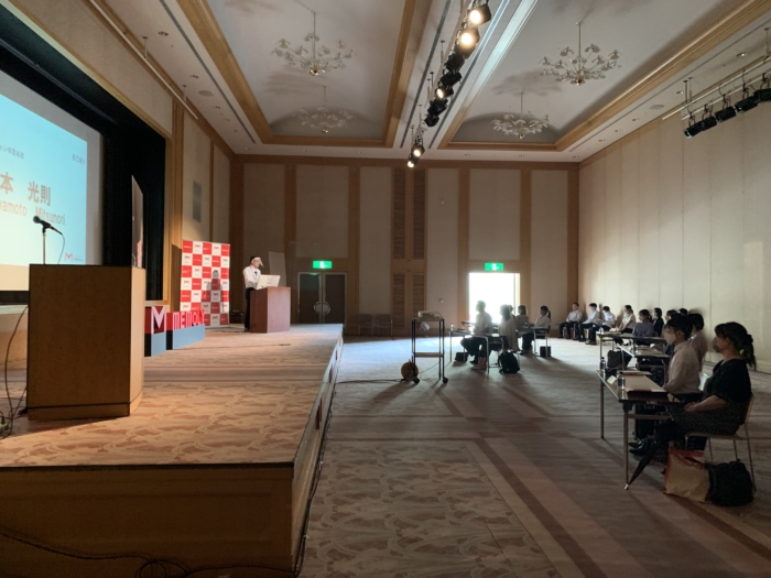 ホールで複数名集まり講演を聞いている